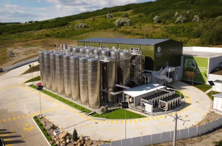 Vedere aeriană asupra cramei Domeniile Averești, cu tancuri de inox, clădire modernă și panouri solare, &icirc;nconjurată de dealuri verzi.