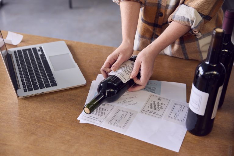Persoană aplic&acirc;nd o etichetă cu cod QR pe o sticlă de vin, l&acirc;ngă un laptop și alte sticle etichetate.