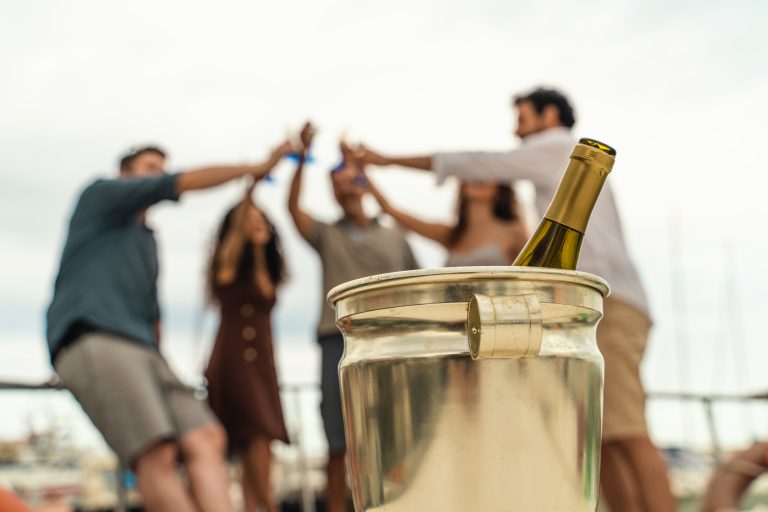 Sticlă de vin alb &icirc;ntr-un recipient cu gheață, &icirc;n prim-plan, cu un grup de prieteni tineri ciocnind &icirc;n fundal
