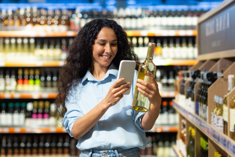 Femeie z&acirc;mbitoare scanează cu telefonul eticheta unei sticle de vin &icirc;ntr-un supermarket.