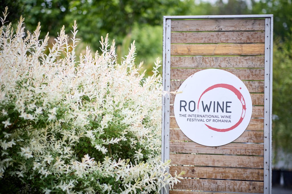 Panou cu sigla RO-Wine &ndash; The International Wine Festival of Romania, amplasat &icirc;n zona de intrare, &icirc;nconjurat de vegetație